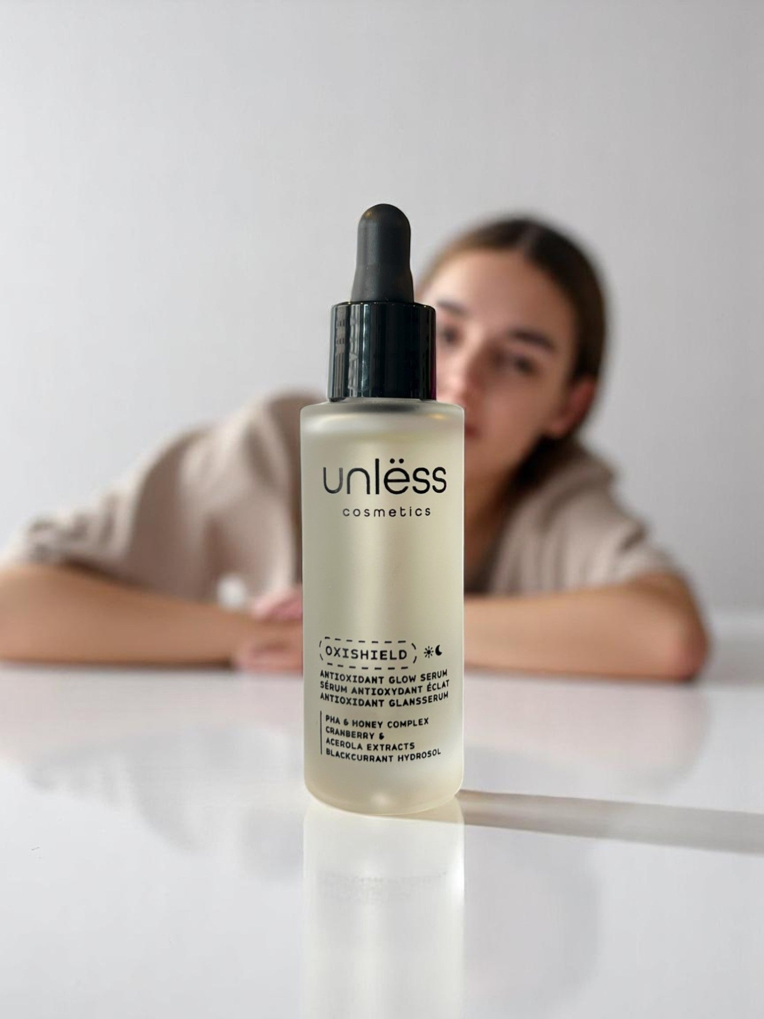 Flacon du sérum Oxishield d’Unlëss Cosmetics vu de face, avec une femme floutée en arrière-plan.