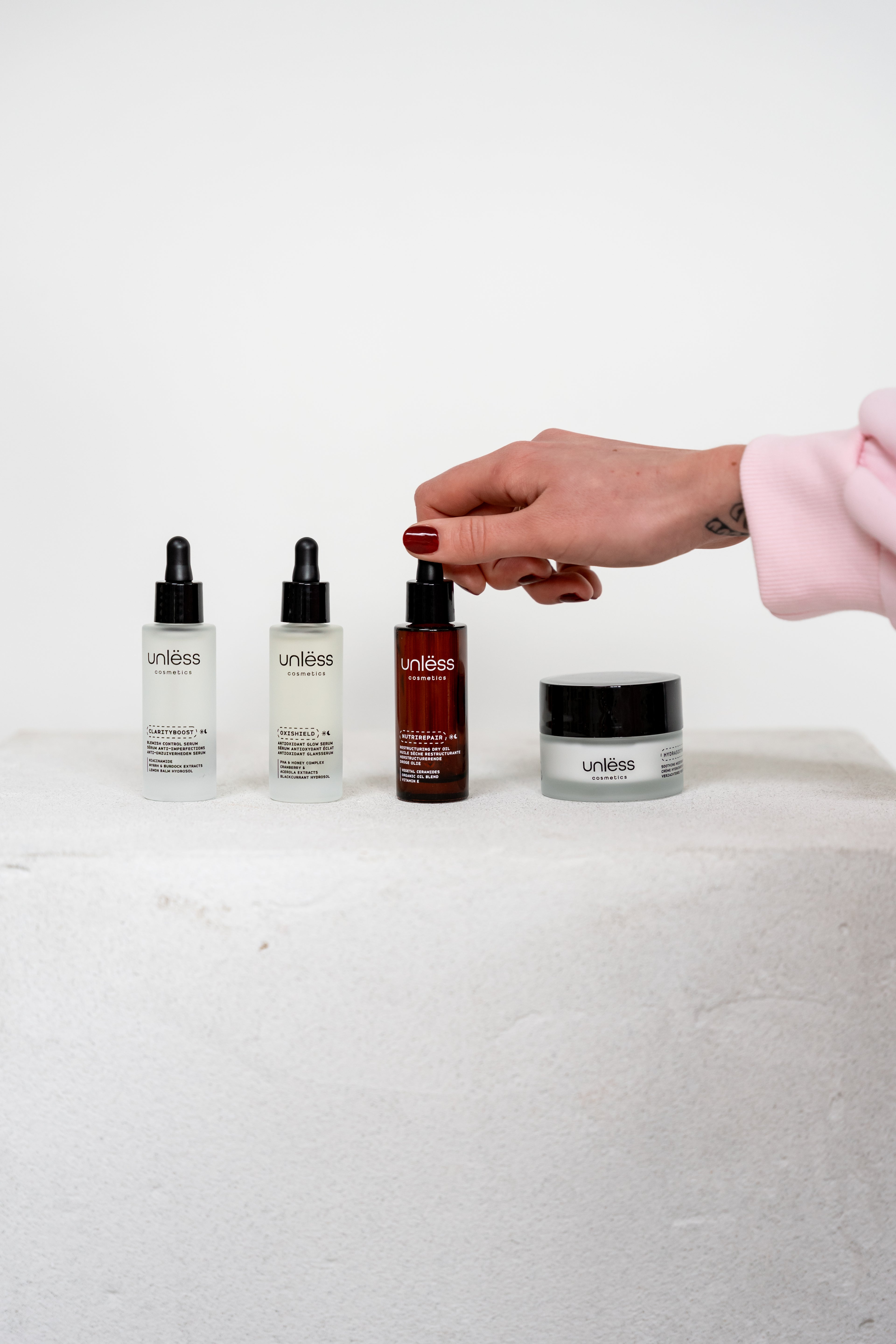 Main saisissant l’huile Nutrirepair d’Unlëss Cosmetics, à côté du sérum Clarityboost, du sérum Oxishield et de la crème Hydrasoothe.