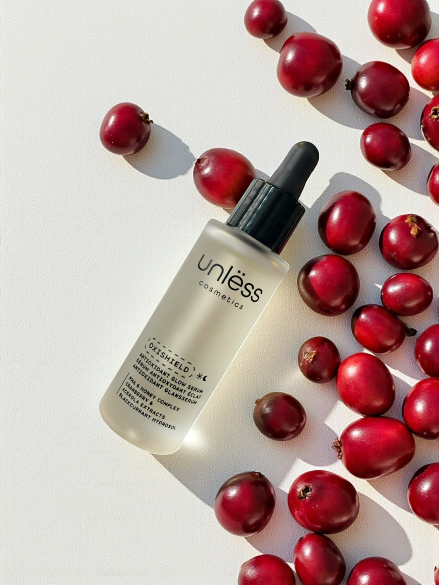 Sérum Oxishield d’Unlëss Cosmetics posé sur un fond texturé blanc, entouré de canneberges rouges avec une ombre marquée du flacon.