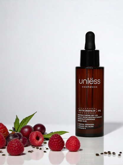 Flacon d’huile posé sur fond blanc, à coté de framboises, de raisin et de graines de cameline.