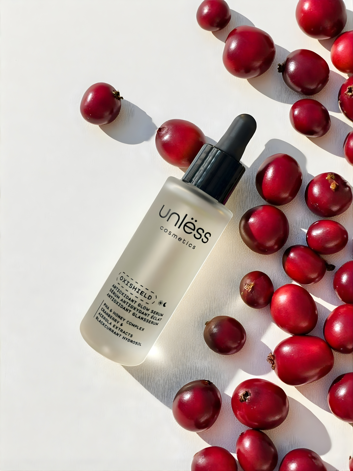 Sérum Oxishield d’Unlëss Cosmetics posé sur un fond texturé blanc, entouré de canneberges rouges avec une ombre marquée du flacon.